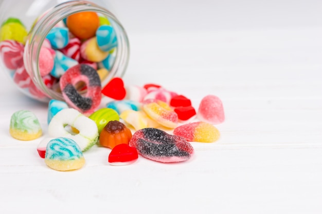 Foto gustosi dolci da masticare colorati e luminosi e caramelle di gelatina cadono dal barattolo sdraiato su una tavola bianca di legno