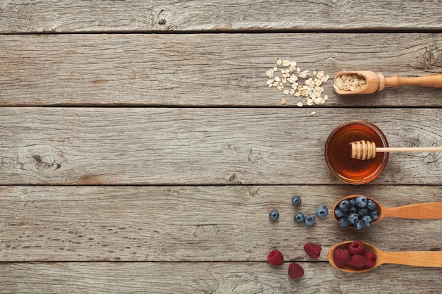 Tasty cooking ingredients background. Blueberries, raspberries, honey, oatmeals in wooden spoons for breakfast on rustic table, top view, copy space, flat lay. Preparing healthy morning meals