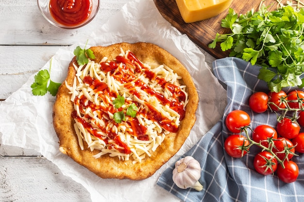 Tasty deep-fried langos served on baking paper with a lot of grated cheese, ketchup, fresh herbs and tomatoes on a checked dish cloth.