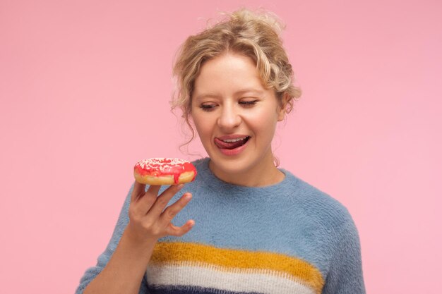Tasty dessert Portrait of woman with short curly hair in sweater holding doughnut looking with desire at donut and licking her lips wants to eat sweet confection indoor studio shot pink background