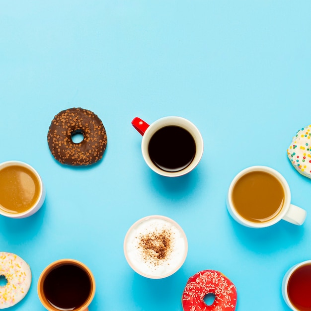 Tasty donuts and cups with hot drinks, coffee, cappuccino, tea on a blue background. Concept of sweets, bakery, pastries, coffee shop, meeting, friends, friendly team. Square. Flat lay, top view.