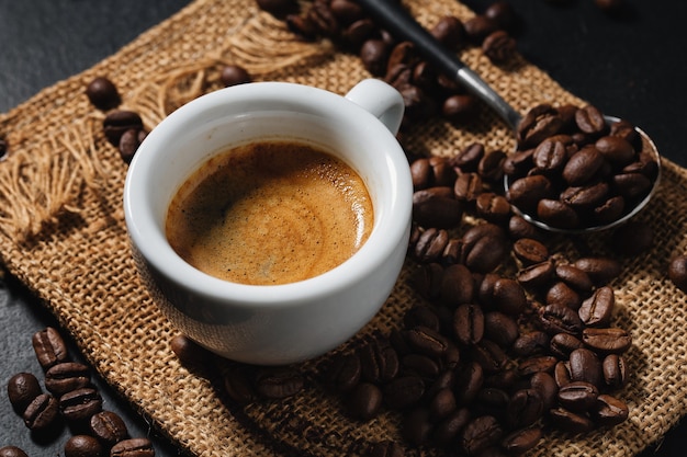 Tasty espresso served in cup with coffee beans around and spoon. Closeup. Dark background.