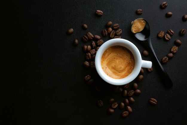 Tasty espresso served in cup with coffee beans around and spoon. View from above. Dark background.