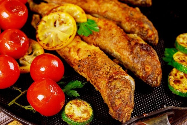 Tasty fish fried with vegetables and lemon on black skillet. Close up.