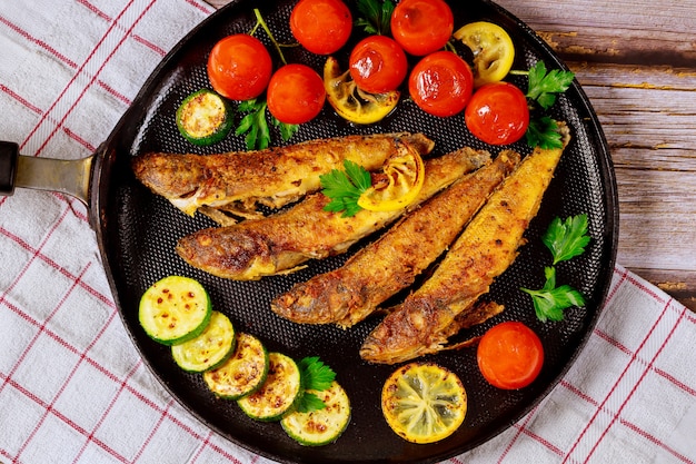 Photo tasty fish fried with vegetables and lemon on black skillet.