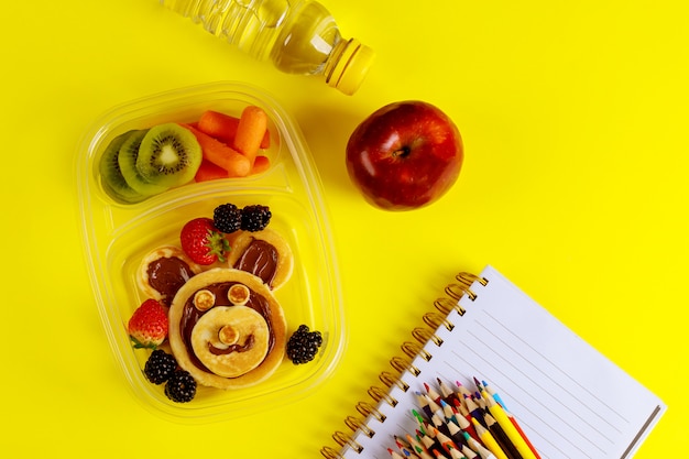 Tasty food in container and colorful pencils on yellow surface