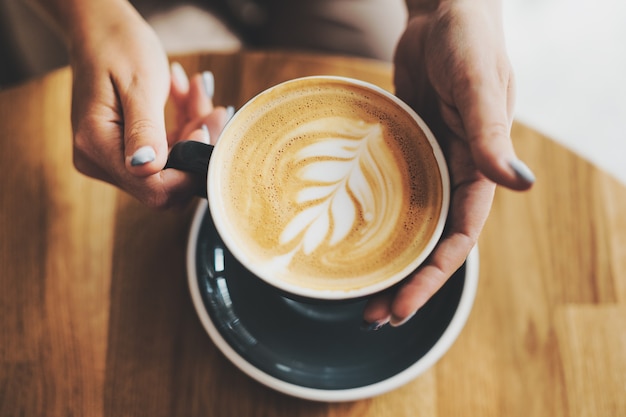 Cappuccino fresco saporito in tazza sulla tavola di legno. donna irriconoscibile che tiene la tazza in mano.