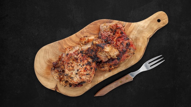 Foto gustosa carne di maiale alla griglia in tagliere di legno d'ulivo naturale accanto alla forcella su tono scuro texture sfondo vista dall'alto piatto