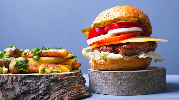 Tasty hamburger with meat cutlet vegetables and cheese with soft bun , fries and champignon mushrooms sprinkled with green onions on forest wooden coasters on a gray background closeup.