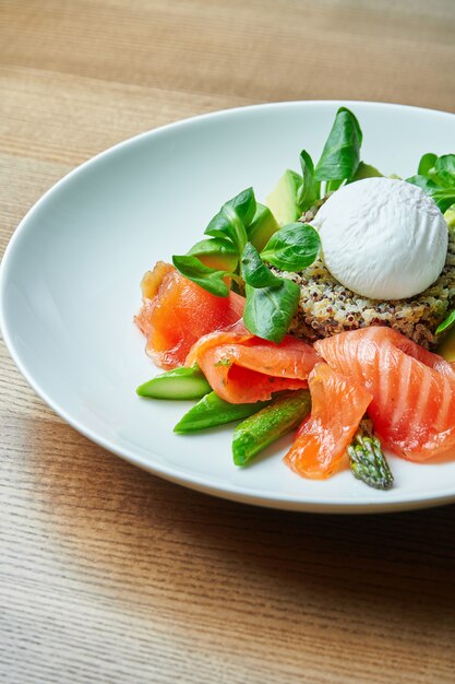 Tasty and healthy continental breakfast - quinoa with benedict eggs, spinach, avocado, salted salmon and asparagus in white bowl. Wooden table