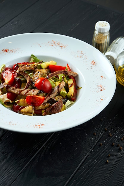 Tasty and healthy warm salad with roast beef and grilled vegetables with olive oil in a white plate on a black wooden surface