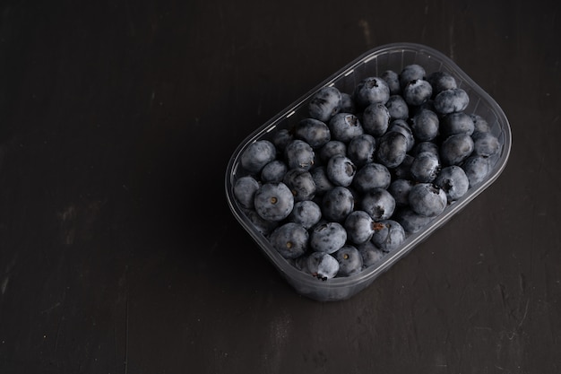 Tasty juicy raw blueberries in a plastic container on a black dark background. Packaging for berries in a supermarket on store shelves.