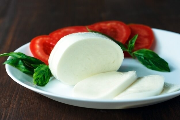 Tasty mozzarella cheese with basil and tomatoes on plate on wooden table