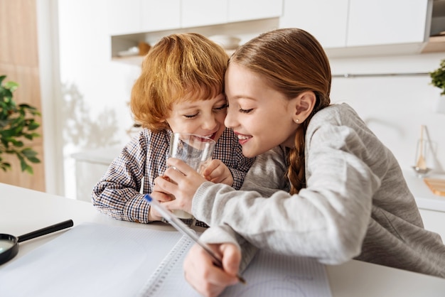 Tasty and nutritious. Beautiful humorous young girl teasing her little brother by trying drinking his milk while working on her home assignment