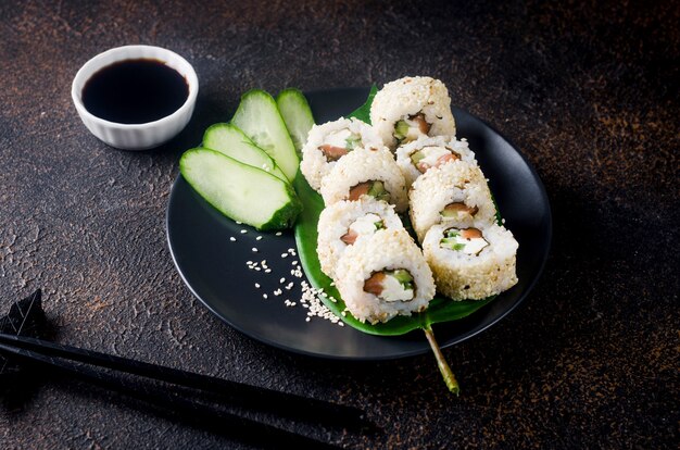 Tasty Philadelphia roll sushi with salmon and cream cheese in sesame on black plate with soy sauce, ginger, wasabi and chopsticks on dark table. Sushi menu. Delivery service Japanese Asian food.