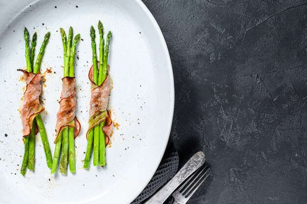 Tasty pork bacon around asparagus in a baking dish. Black background. Top view. Copy space