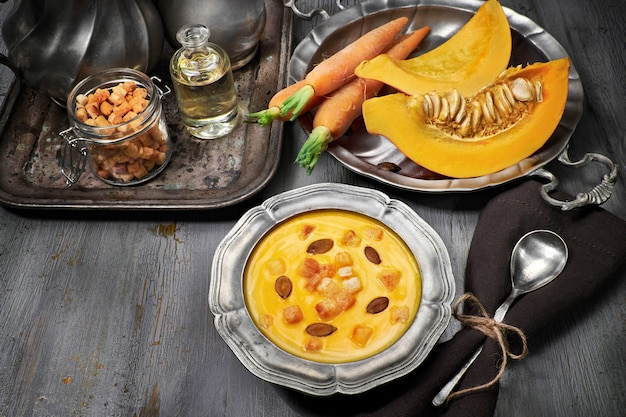 Tasty pumpkin and carrot cream soup served with croutons in metal pewter bowl with ingredients
