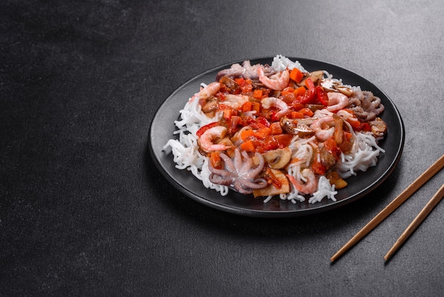 Tasty rice noodles with tomato, red pepper, mushrooms and seafood on a dark concrete background