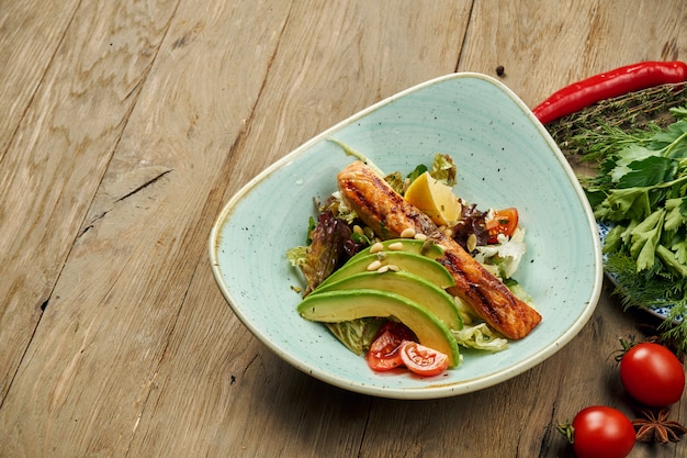 Tasty salad with grilled salmon, avocado, lettuce in a blue bowl on a wooden surface. Seafood salad