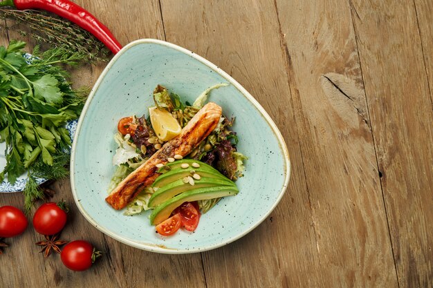 Tasty salad with grilled salmon, avocado, lettuce in a blue bowl on a wooden surface. Seafood salad