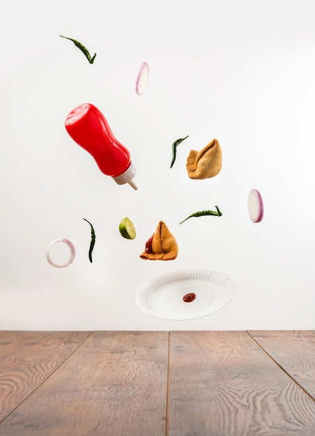 Tasty Samosa Snacks flying with ketchup bottle, green chilli, onion slices and plate over plain background