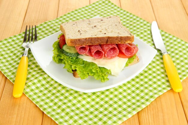 Tasty sandwich with salami sausage and vegetables on white plate on wooden background