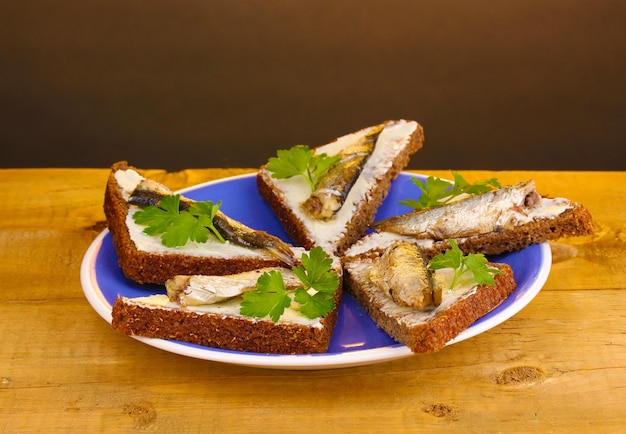 Tasty sandwiches with sprats on plate on wooden table on brown background