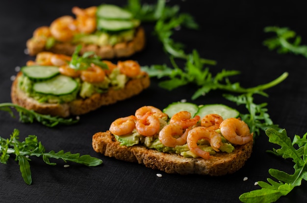 Tasty snack of toasts with avocado, fried shrimps, cucumber and arugula