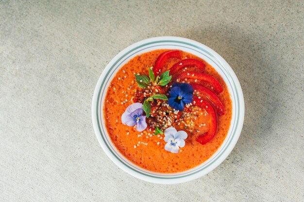 Tasty tomatoe soup in white bowl, decorated with little flowers, red bell pepper, hempseed and mint.