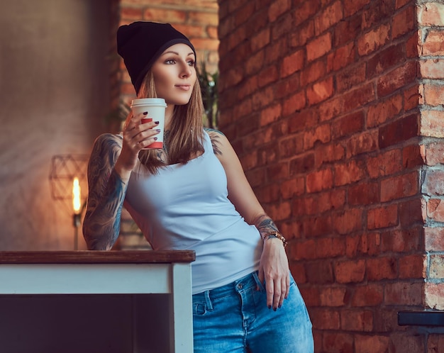 A tattoed sexy blonde in a t-shirt and a hat with cup of coffee in a room with loft interior.