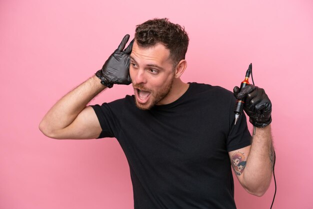 Tattoo artist brazilian man isolated on pink background listening to something by putting hand on the ear