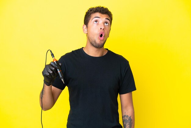 Tattoo artist Brazilian man isolated on yellow background looking up and with surprised expression