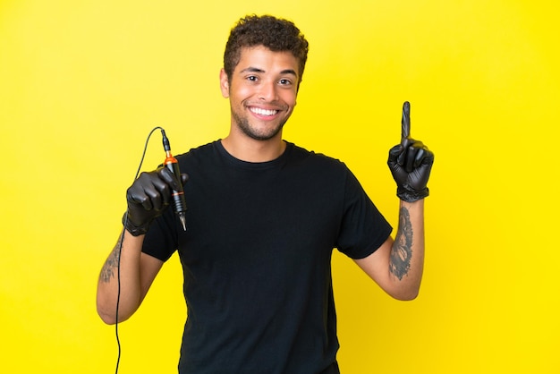 Tattoo artist Brazilian man isolated on yellow background showing and lifting a finger in sign of the best