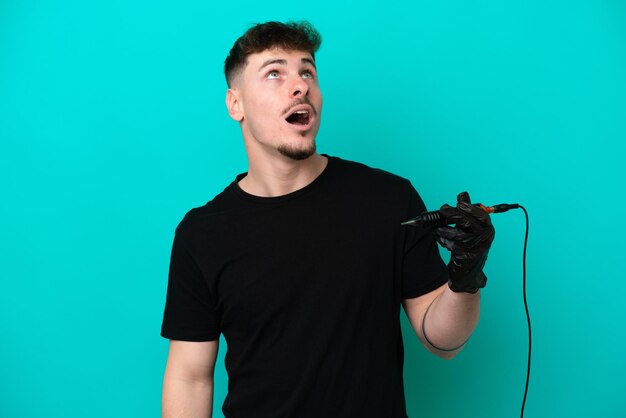 Tattoo artist caucasian man isolated on blue background looking up and with surprised expression