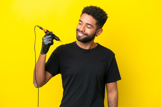 Tattoo artist man Brazilian man isolated on yellow background looking to the side and smiling