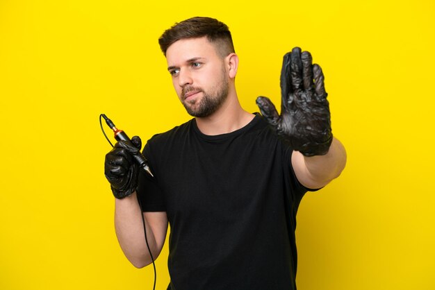 Tattoo artist man isolated on yellow background making stop gesture and disappointed