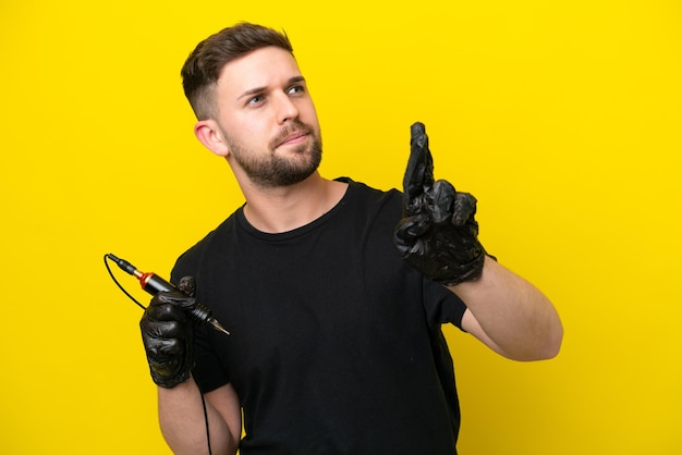 Tattoo artist man isolated on yellow background with fingers crossing and wishing the best