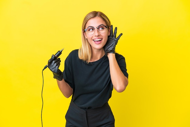 Tattoo artist uruguayan woman isolated on yellow background listening to something by putting hand on the ear