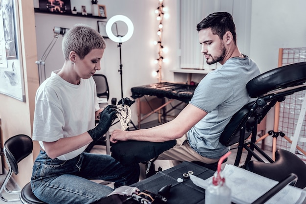 Maestro del tatuaggio che lavora. attenta donna attenta che attacca schizzo speciale con l'immagine del futuro tatuaggio su di esso sulla mano del cliente