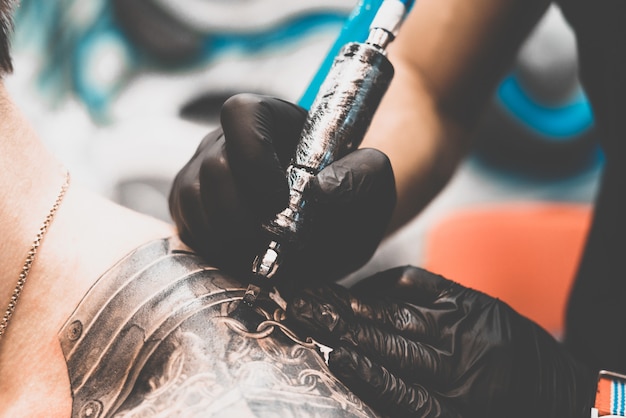 Tattoo salon. The tattoo master is tattooing a man on his shoulder. Tattoo machine, safety and hygiene at work. Close-up, tinted, tattooist