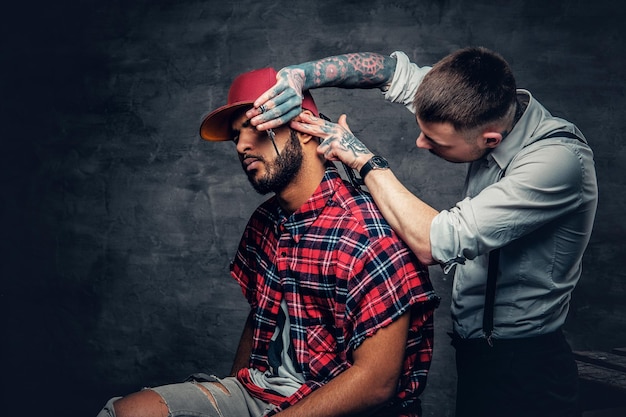 Barbiere tatuato che taglia la barba di un maschio hipster nero con un berretto da baseball.