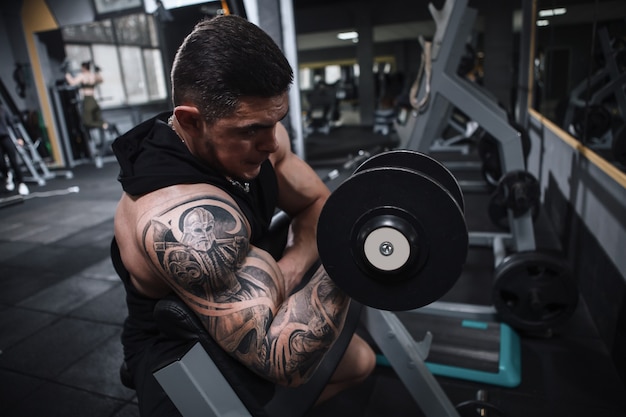 Татуированный культурист делает сгибания бицепса в спортивной студии
