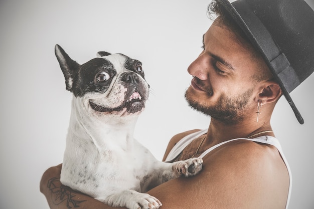 Ragazzo tatuato con piercing con cane bulldog francese tra le braccia su sfondo bianco