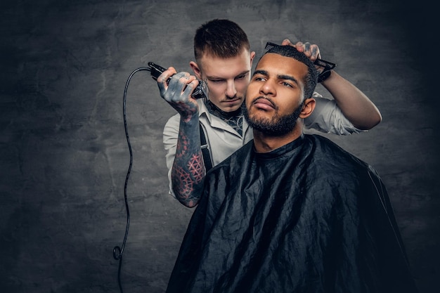 Tattooed Caucasian male barber cutting the beard to Black stylish man.