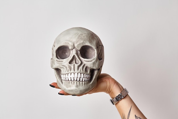 검은색 시계에 문신을 한 여성의 손이 흰색으로 격리된 이빨을 가진 인간 두개골의 현실적인 모델을 들고 있습니다. 의학 또는 할로윈 공포 개념 클로즈업 샷