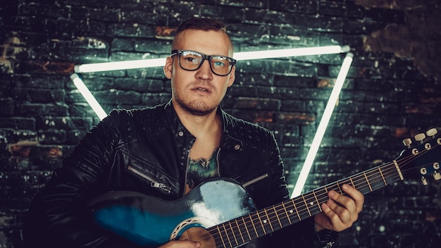 Татуированный мужчина играет на гитаре возле освещенной стены