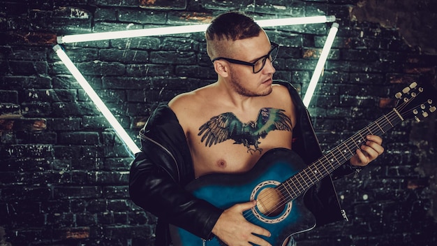 Tattooed man playing guitar near illuminated wall stylish guy\
with bird tattoo on chest looking away and playing guitar while\
standing against shabby brick wall with triangle illumination