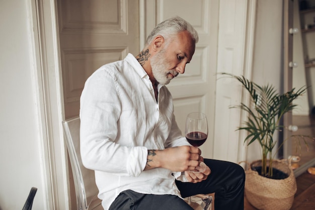 Uomo tatuato in camicia bianca moderna con bicchiere di vino bel ragazzo adulto triste con barba grigia con bevanda rossa nelle sue mani