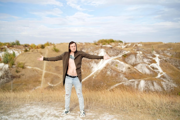 Tattooed man with bare torso in cape and jeans stands in hilly terrain Adult male poses on cloudy day in countryside