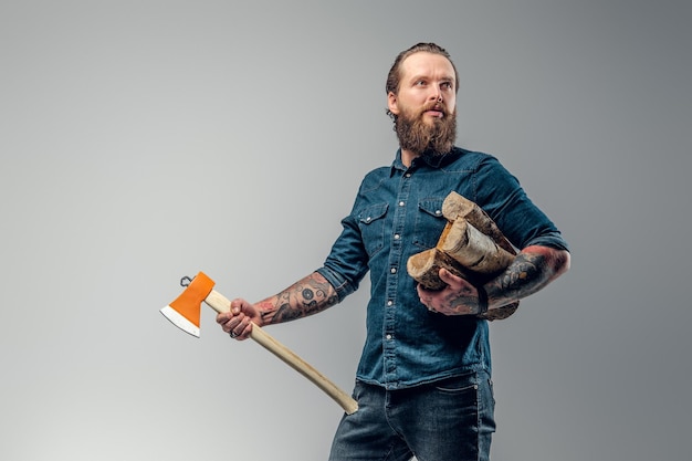 Photo tattooed man with firewood and axe in hands is posing for photographer.